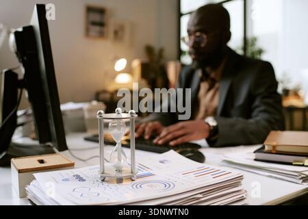 Black Man Working at Desk with Hourglass in Modern Office Stock Photo