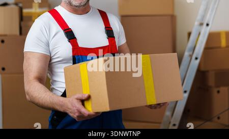 Man carries a brown box in a storage area with many boxes stacked Stock Photo