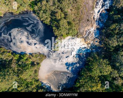 A vertical aerial view shows swirling foam patterns in a dark river pool, bordered by dense forest and rocky terrain. Stock Photo