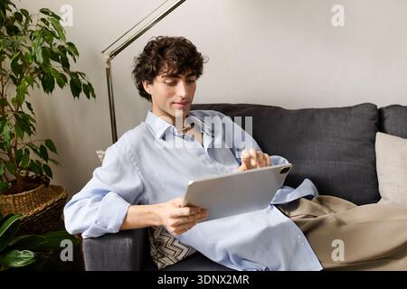Curly-haired young man relaxes in his stylish apartment while engaging with his tablet. Stock Photo