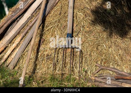 Rustic farm pitchfork leaning on a haystack with wooden poles nearby, sunlit rural scene, harvest and agriculture concept. Stock Photo