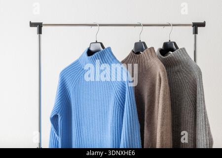 Soft textured sweaters of blue, taupe and grey luxury natural cashmere wool hanging on metallic hanger against white wall background. Stock Photo