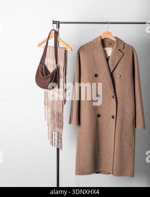 Brown wool  double breasted coat or jacket, beige alpaca scarf and dark suede handbag on hanger against light grey wall background. Warm wool clothing Stock Photo