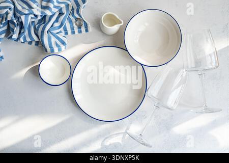 Top view of a table setting featuring white dishware with blue accents, two empty wine glasses, and a striped blue and white cloth on a light textured Stock Photo