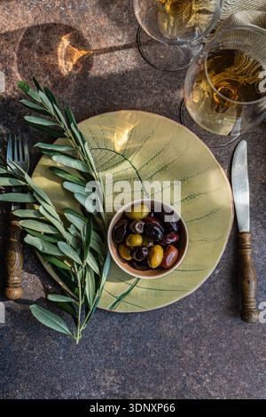 Top view of a table set with Greek olives in a bowl on a yellow plate with leaf pattern, accompanied by amber wine in glasses, on a textured stone sur Stock Photo