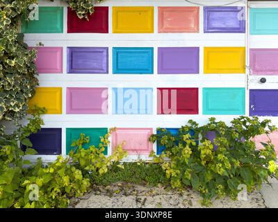An old white door is adorned with vibrant, colorful rectangular panels in various shades. Surrounding the door, green foliage adds a natural touch to Stock Photo