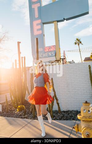 A woman in a red dress and white boots stands by a vintage Las Vegas motel sign, surrounded by desert cacti, with the sun setting behind her, casting Stock Photo