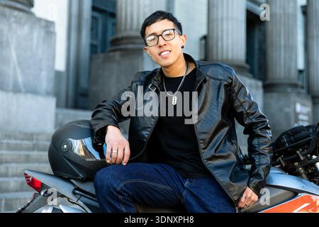 A Hispanic man, stylishly dressed in a black leather jacket, sits confidently on his motorcycle in an urban setting, showcasing a modern urban lifesty Stock Photo