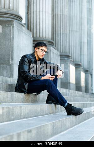 Hispanic man in a black leather jacket and jeans sits on stone steps outside, focused on his smartphone. The setting features tall stone columns, sugg Stock Photo