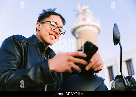 Hispanic male motorcyclist in a leather jacket standing near a city lighthouse. He is holding a smartphone, smiling, and enjoying his urban adventure. Stock Photo