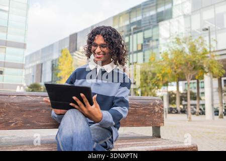 A non-binary individual with curly hair, smiling while using a tablet outdoors. The urban background and casual attire emphasize a modern lifestyle an Stock Photo
