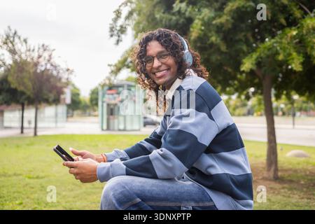 A joyful non-binary person with headphones and a phone relaxes in an urban park. Embracing modern LGBT lifestyle, this candid city scene highlights di Stock Photo
