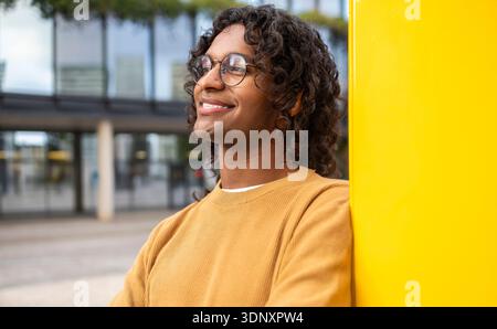 Non-binary individual with curly hair and glasses stands confidently against a yellow backdrop in an urban setting, showcasing vibrant city lifestyle Stock Photo