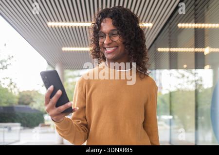 A non-binary individual with curly hair and glasses smiles warmly while using a smartphone under an urban canopy, reflecting a modern and inclusive li Stock Photo