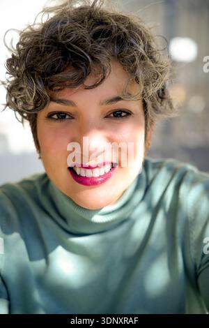 Close-up portrait of a woman with short curly hair, wearing a green sweater. She smiles warmly at the camera with bright pink lipstick, under soft nat Stock Photo