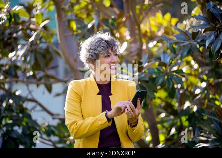A confident businesswoman in a vibrant yellow jacket is happily engaging with her smartphone outdoors. Her surroundings are lush and green, creating a Stock Photo