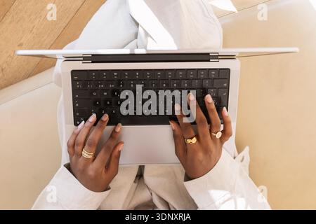 Overhead view of a black Gen Z woman typing on a laptop, showing her hands with rings on her fingers. She is seated on a light colored leather couch i Stock Photo