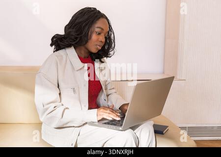 A young Black Gen Z woman sitting on a sofa, working intently on her laptop. She wears a light jacket over a red sweater, representing modern work fro Stock Photo