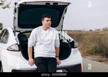 A young man wearing a white sweatshirt and black pants sits on the back of an electric car with an open trunk. The setting is outdoors along a quiet r Stock Photo