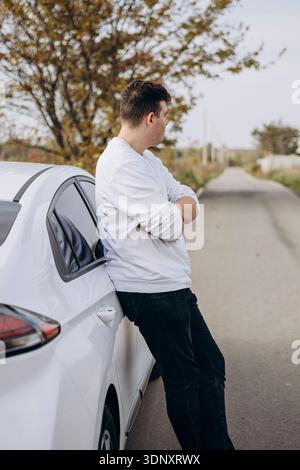 A young man in casual attire leans against a sleek electric car on a quiet countryside road, contemplating the scenic view under the autumn sky. Stock Photo