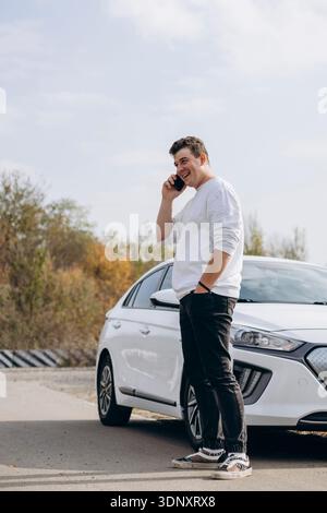 A young man stands next to his electric car while talking on the phone outdoors. He appears relaxed and content, enjoying a moment amidst a peaceful b Stock Photo