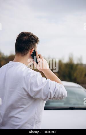 A young man in a white shirt stands by his electric car, making a phone call. The setting is outdoors, with a scenic background, symbolizing modern co Stock Photo