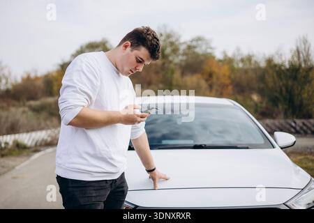 A young man stands by his parked electric car on a scenic road, engrossed in his smartphone. The scene captures modern technology and sustainable trav Stock Photo