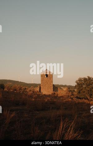 A solitary stone tower stands amidst burnt grass at sunset. In the distance, wind turbines dot the hill, silhouetted against the orange sky, blending Stock Photo
