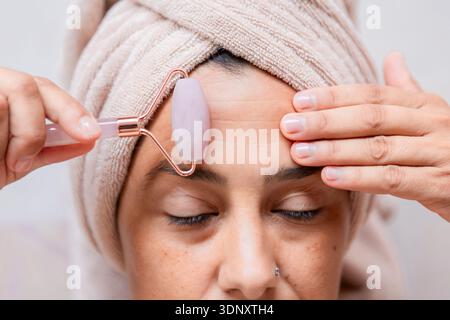 A woman with a towel wrapped around her head uses a rose quartz jade roller on her forehead, embodying relaxation and skincare. The tool promotes beau Stock Photo