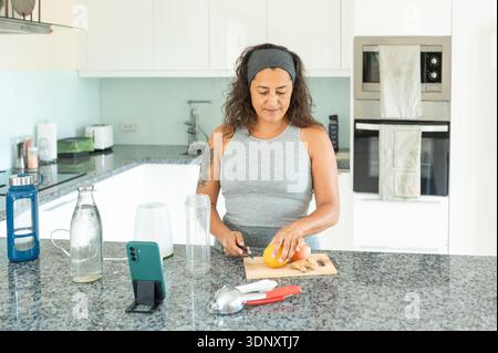 Healthy woman preparing fresh fruit juice in her modern kitchen. She slices an orange on a cutting board while using a smartphone for a recipe. Blende Stock Photo