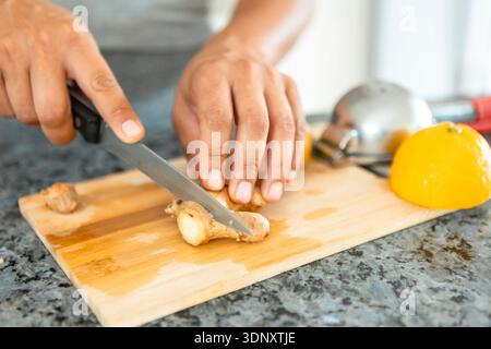 Cropped unrecognizable woman slicing ginger on a wooden board beside a lemon, emphasizing healthy meal preparation at home with fresh ingredients. Stock Photo