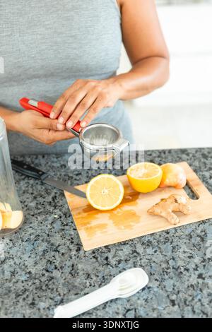 Cropped unrecognizable woman squeezing fresh lemon juice with a handheld juicer on a kitchen counter. Sliced ginger and lemon are on a wooden board. Stock Photo