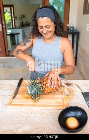 Smiling woman slicing a ripe pineapple on a wooden board in a cozy home kitchen setting. She appears focused and content, engaging in healthy meal pre Stock Photo