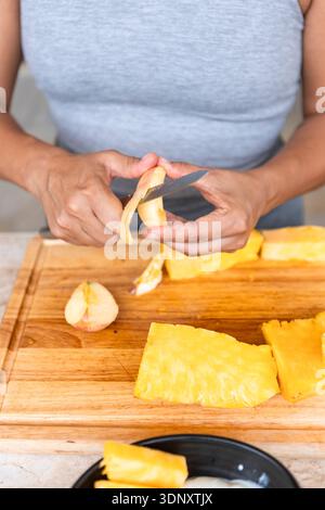 Cropped unrecognizable woman slicing fruits on a wooden cutting board. She is carefully peeling and preparing healthy snacks in a home kitchen setting Stock Photo