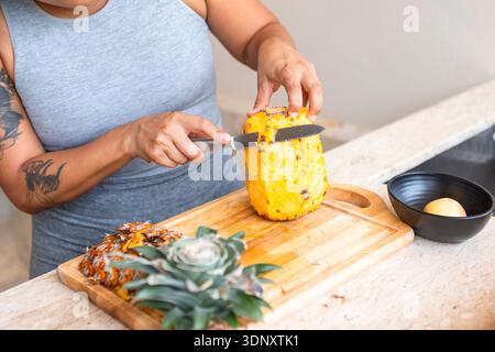 Cropped unrecognizable woman slicing a fresh pineapple on a wooden chopping board at home, demonstrating a healthy home routine and promoting wellness Stock Photo