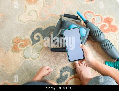 Top view of a cropped unrecognizable woman's hand holding a smartphone while standing on a colorful carpet. Nearby, fitness equipment lies on the floo Stock Photo