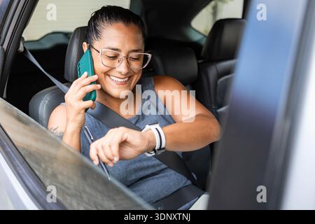 A businesswoman is seated in a car, engaging in a call on her smartphone while checking her smartwatch. She embodies productivity and multitasking in Stock Photo