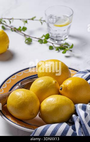 A collection of fresh lemons sits in a decorative yellow and blue plate, accompanied by a wooden spoon and striped cloth. A sprig and glass of water c Stock Photo