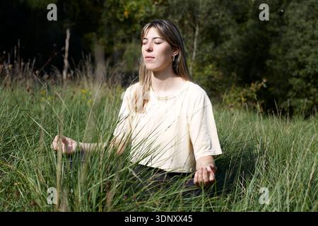 Woman sitting peacefully in a forest, meditating and connecting with nature. The tranquil environment enhances mindfulness and relaxation, promoting a Stock Photo
