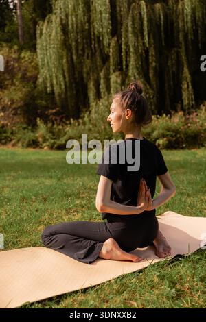 Young woman practicing yoga outdoors on a sunny day, seated on a mat in a serene park. She sits cross-legged, focusing on mindfulness and the natural Stock Photo