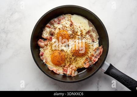 Top view of sizzling fried eggs accompanied by crispy bacon in a black skillet on a sleek white marble surface. Perfect savory breakfast scene. Stock Photo
