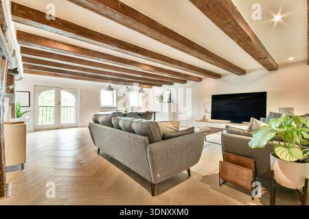 A spacious modern living room with exposed wooden beams and herringbone flooring. Features a grey sofa, large TV, and big windows allowing natural lig Stock Photo