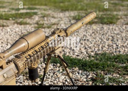 A close-up view of an airsoft sniper rifle in camouflage on a training field. The weapon is set on a bipod, highlighting its realistic design and tact Stock Photo