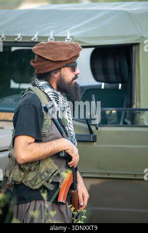 A bearded man wearing tactical military gear stands near a vintage army vehicle. He holds a replica firearm, embodying the intensity of airsoft battle Stock Photo