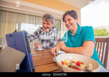 Transgender couple sharing a joyful breakfast and engaging in lively conversation while using a tablet at an outdoor setting, capturing a moment of co Stock Photo