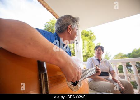 Two individuals enjoy a relaxing morning on a porch. One is playing the guitar while the other listens, creating a cozy and cheerful atmosphere in a h Stock Photo
