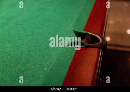 A detailed close-up view of a pool table, highlighting the vibrant green felt and sturdy pocket. The wood finish and lighting create a warm, inviting Stock Photo