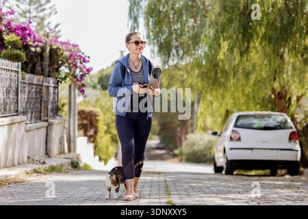 A woman walks along a tree-lined street, holding a yoga mat and using her mobile phone. A cat follows closely beside her, creating a peaceful outdoor Stock Photo