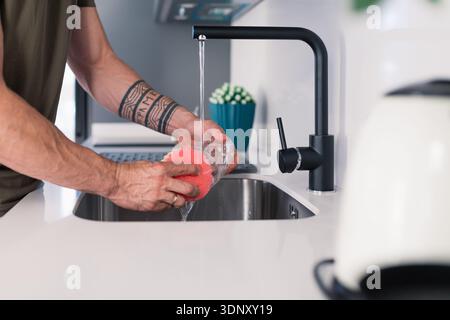 A man cleans a glass with a red sponge under a sleek black faucet in a modern kitchen with white countertops, showcasing everyday home life and cleanl Stock Photo