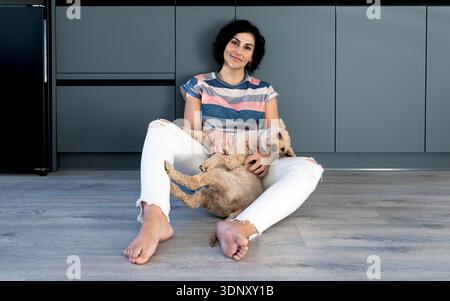 A woman sits comfortably on a kitchen floor, joyfully holding her dog. The atmosphere is warm and cozy, highlighting the bond between the woman and he Stock Photo